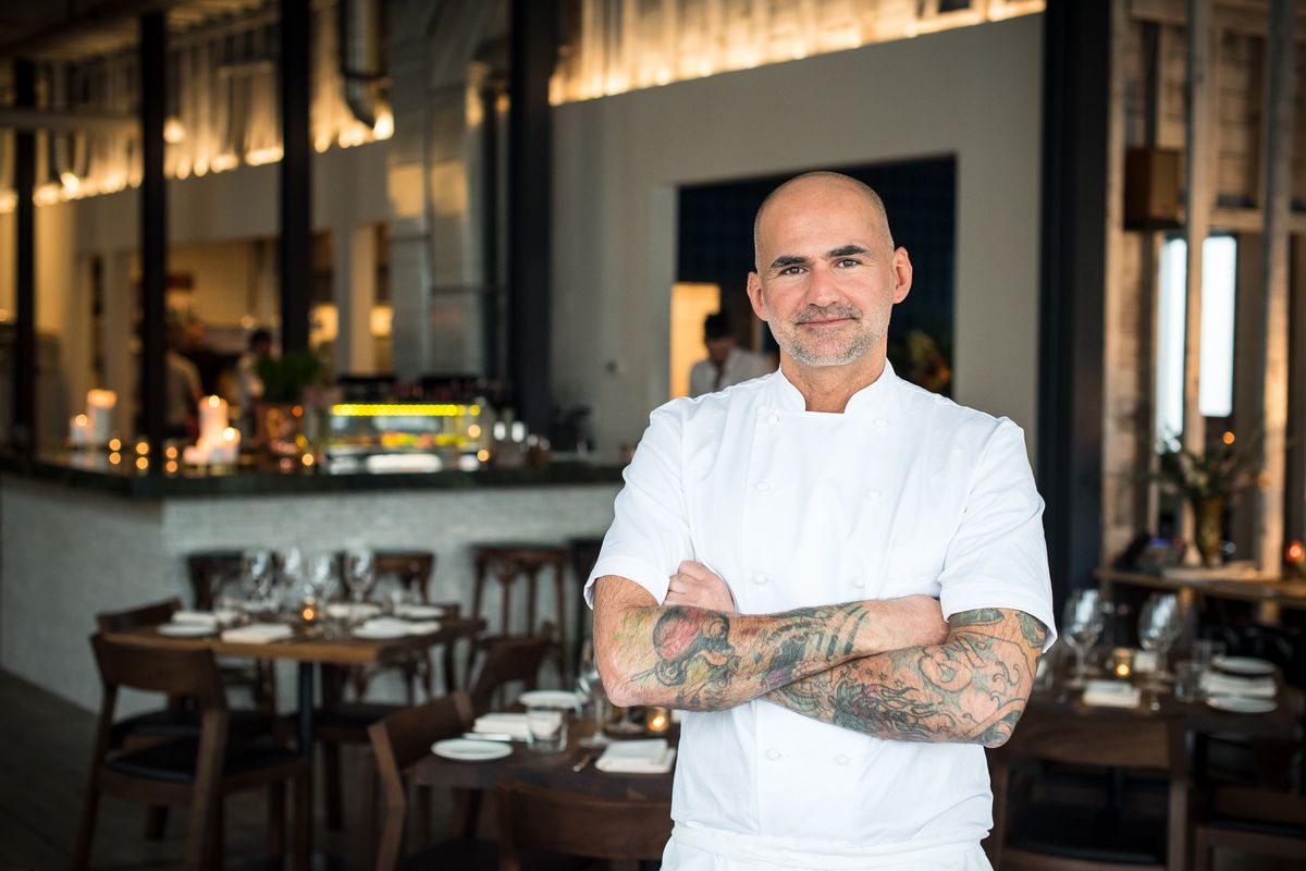 The bald, tattooed chef with a salt and pepper goatee stands, arms crossed, inside a dining room with wood tables and white walls