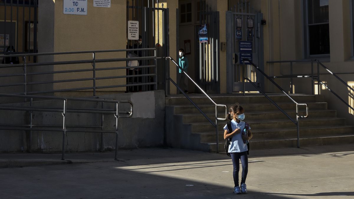 A young girl, wearing a blue shirt and protective mask, waits outside of school for her parent. Another young student stands in the background next to the entrance of the school.