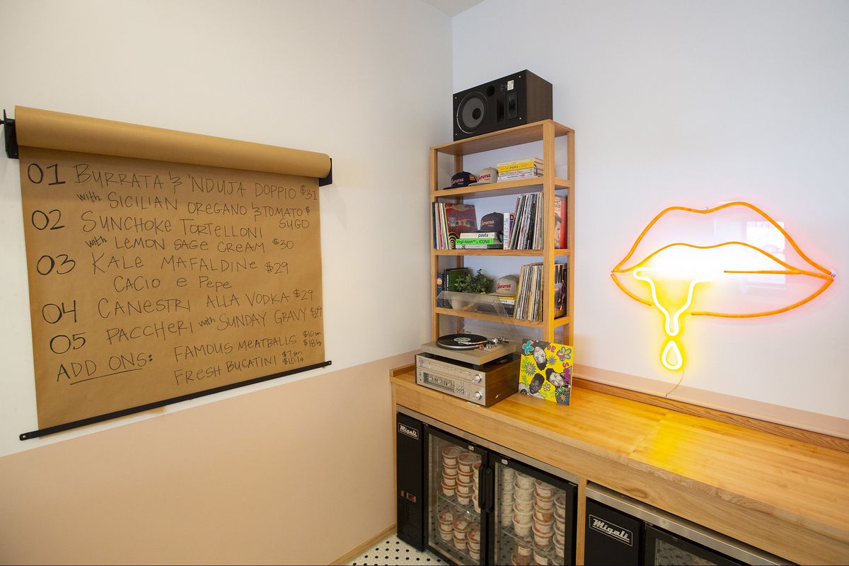 A hand-written menu on butcher paper hangs on the wall beside a countertop upon which sit a stereo and a bookcase, and below, a minifridge filled with plastic food containers. On the wall above the counter, a neon sign of a drooling mouth.