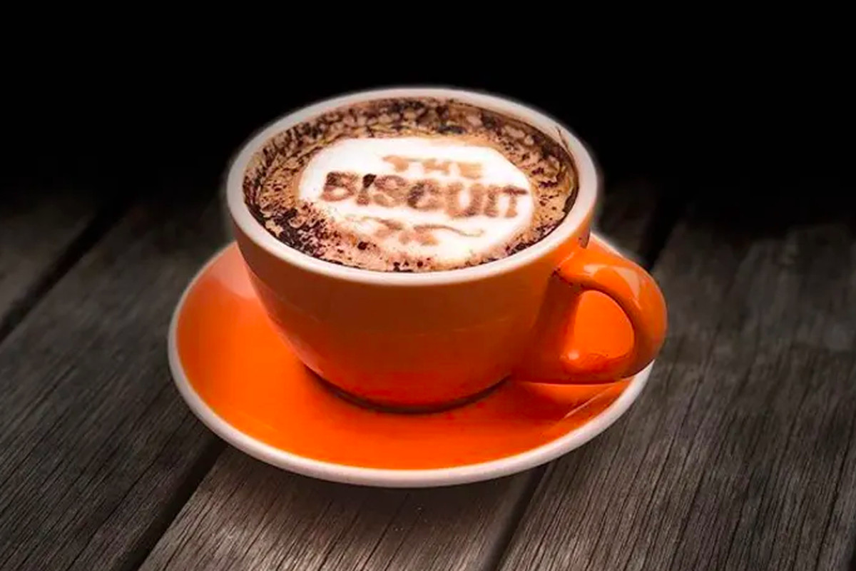 A latte in an orange mug has “the biscuit” written in the foam