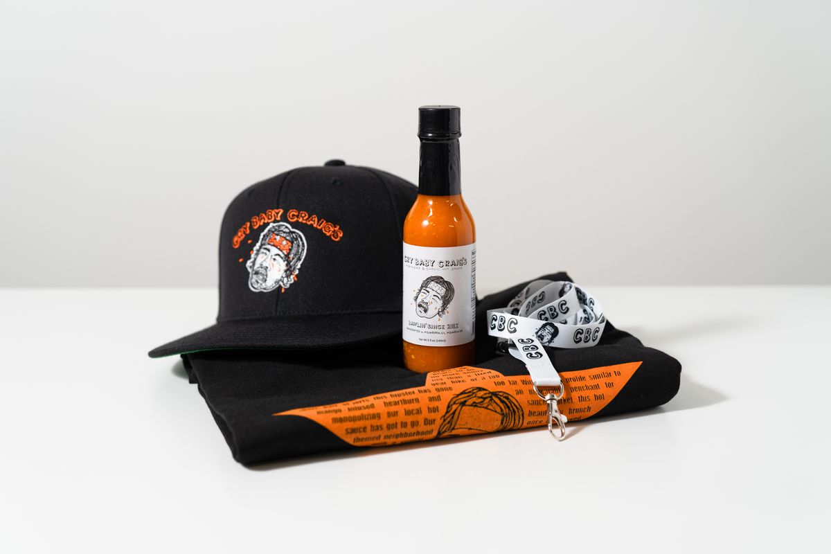 A folded t-shirt, bottle of hot sauce, lanyard, and hat from Cry Baby Craig