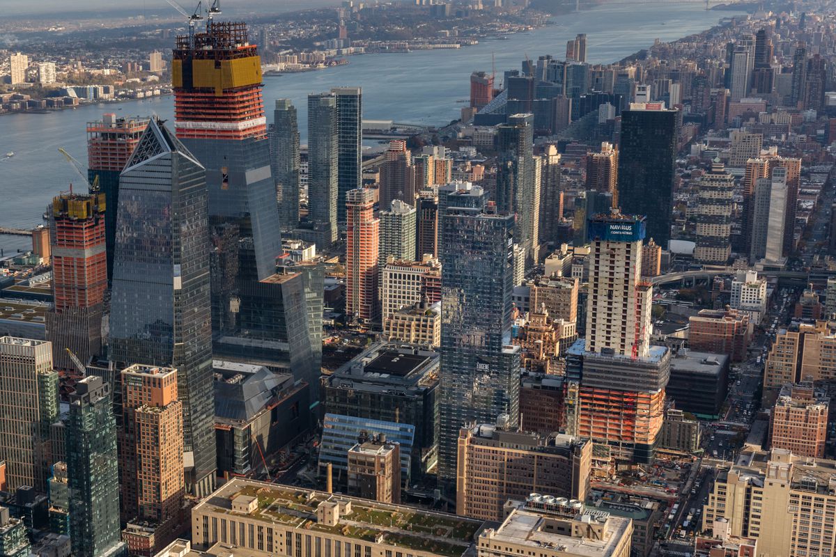 An aerial view of Hudson Yards in New York City. There are many tall city buildings next to each other. There is a body of water in the distance.