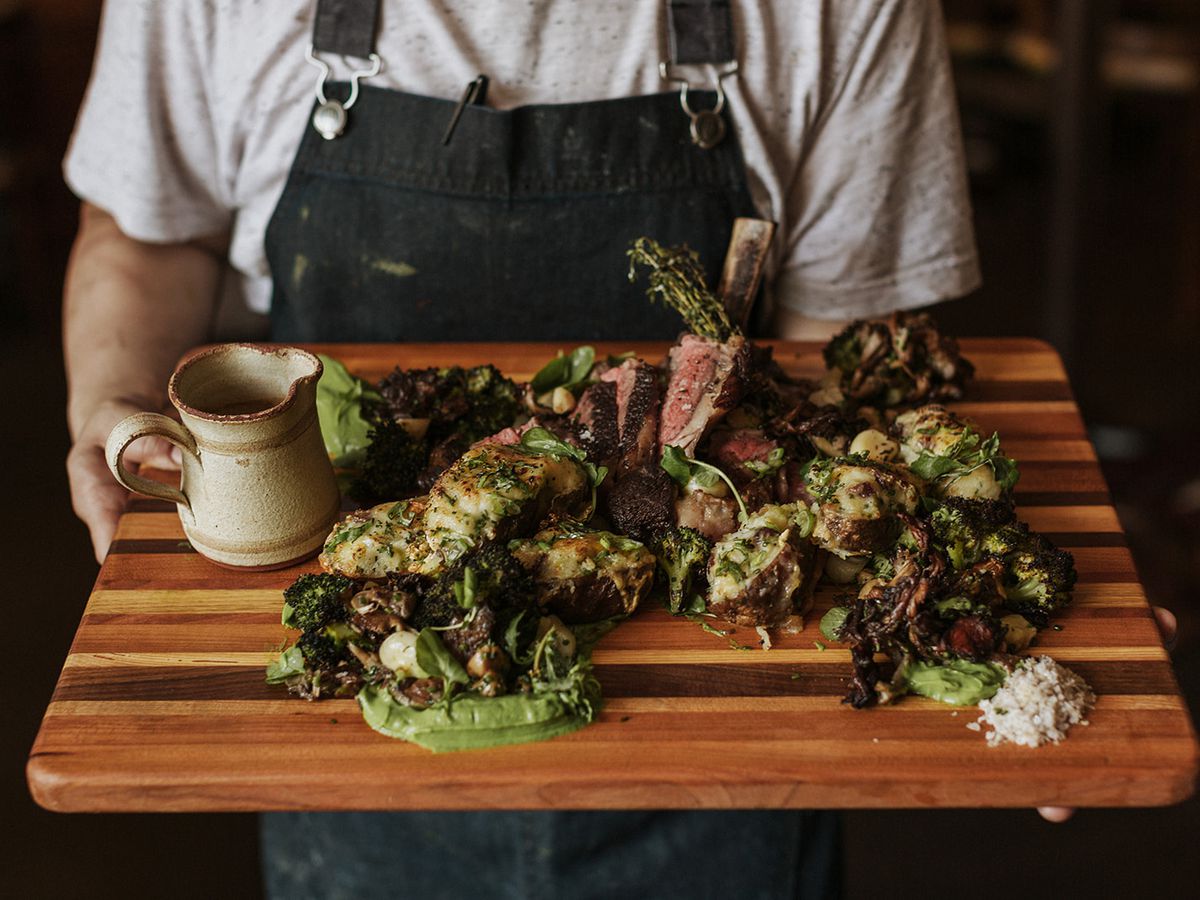 A server holds a large wooden board covered with sliced Côte de Boeuf doused in green sauce with a small pitcher to one side