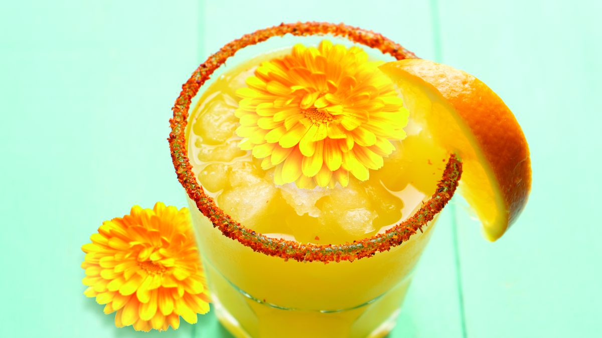 The Don Francisco Margarita, in a rocks glass, topped with a marigold and garnished with a salt rim and orange slice. The cocktail glass is sitting next to another marigold on a wooden table.