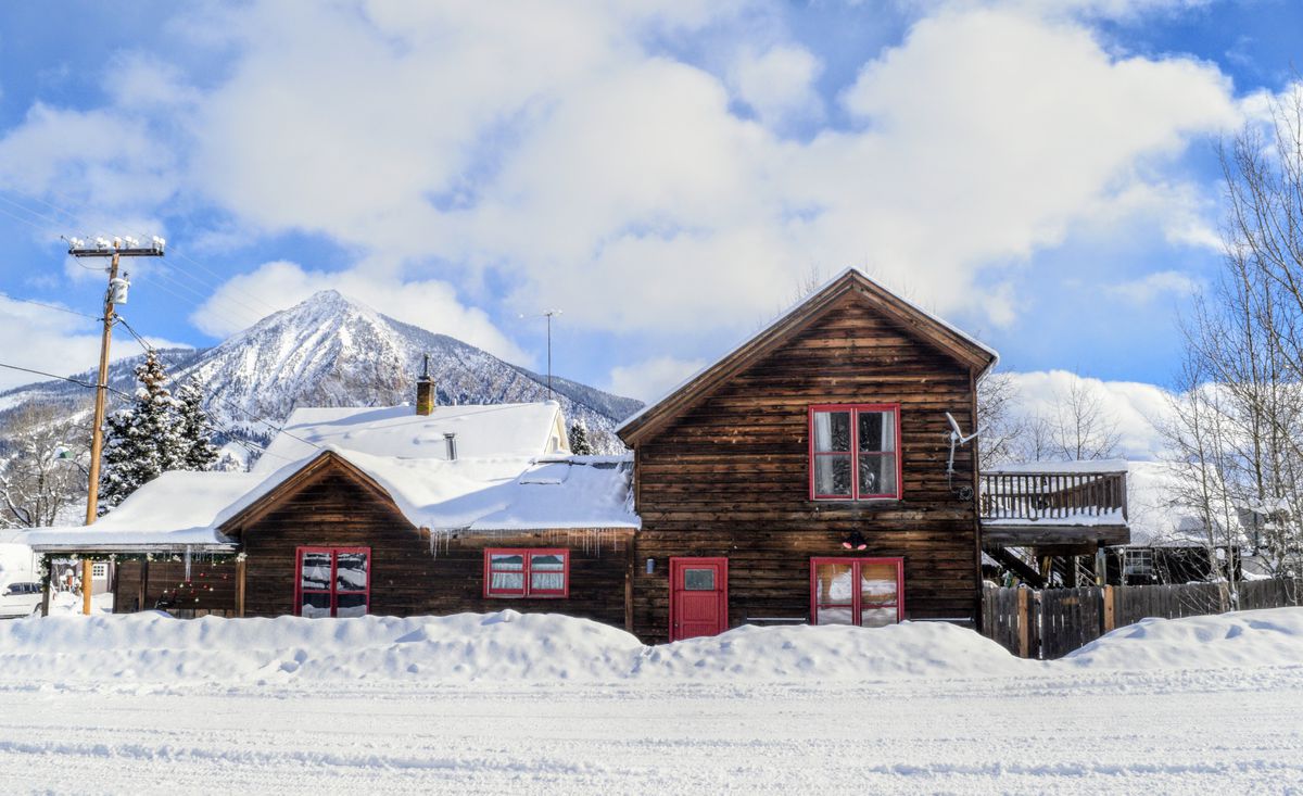 A rustic wooden home with red trim and covered in snow sits behind a snowy street. In the background you can see a snow-capped mountain, blue skies, and fluffy white clouds.