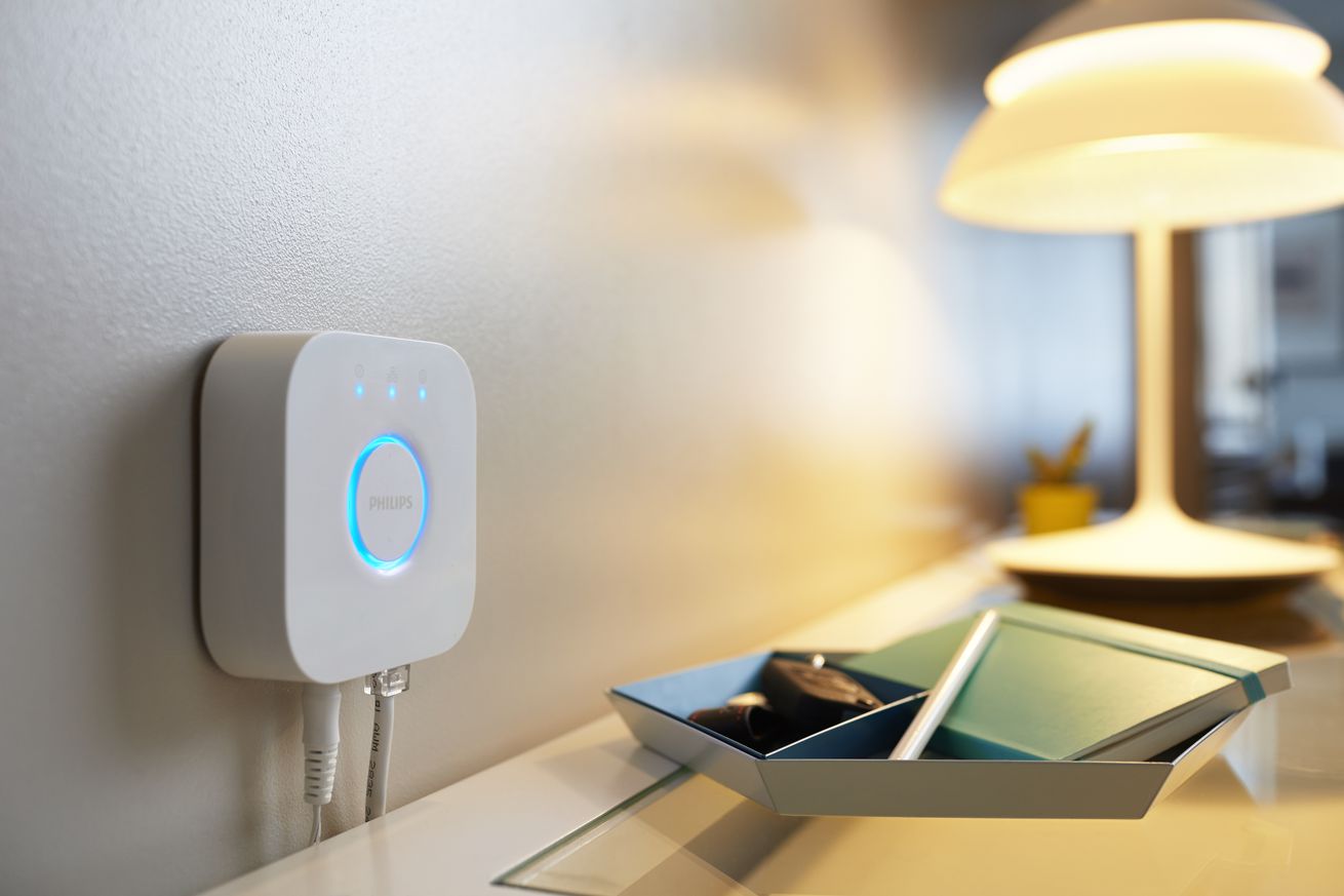 Philips Hue Bridge device shown wall-mounted above a table, near a lamp.