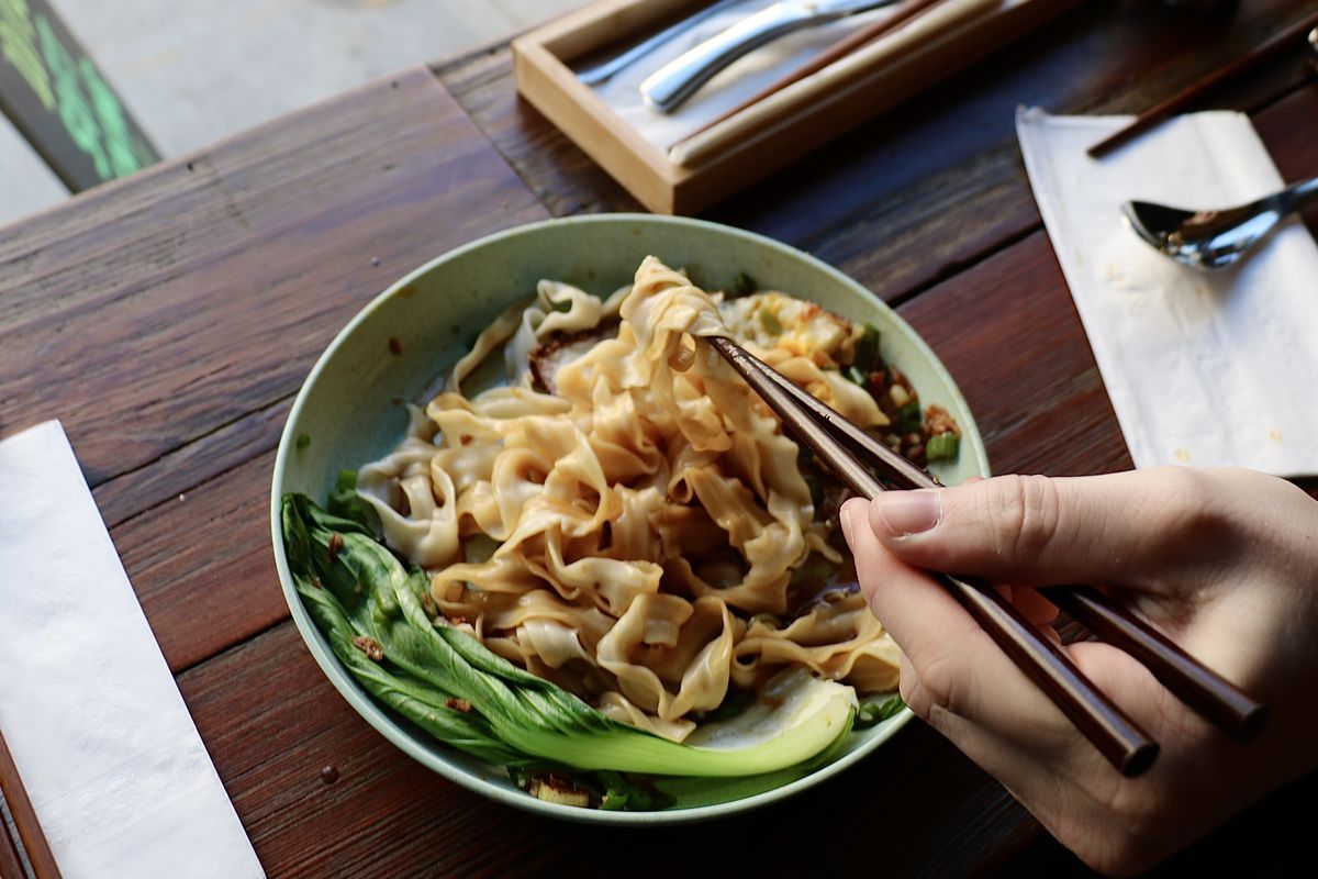 A hand holding chopsticks pulls a noodle from a bowl of scallion noodles.