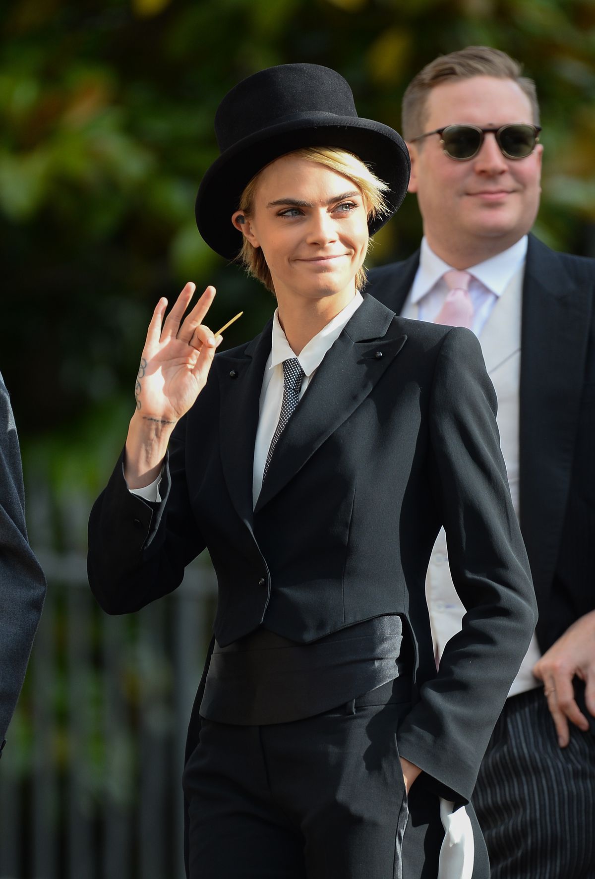 Cara Delevingne waves to someone, toothpick in hand.