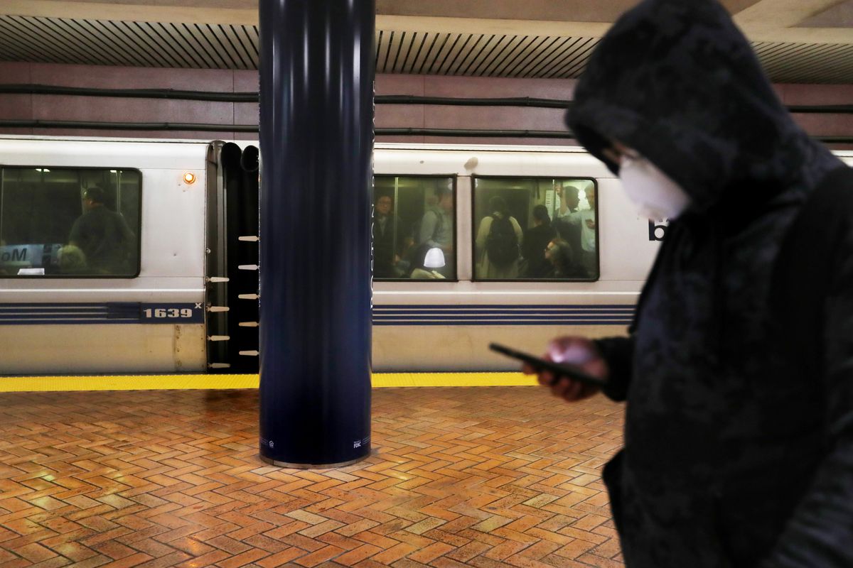 A man wearing a dark hoodie and a white mask over his nose and mouth, while standing on a train platform with a blue and gray train behind him.