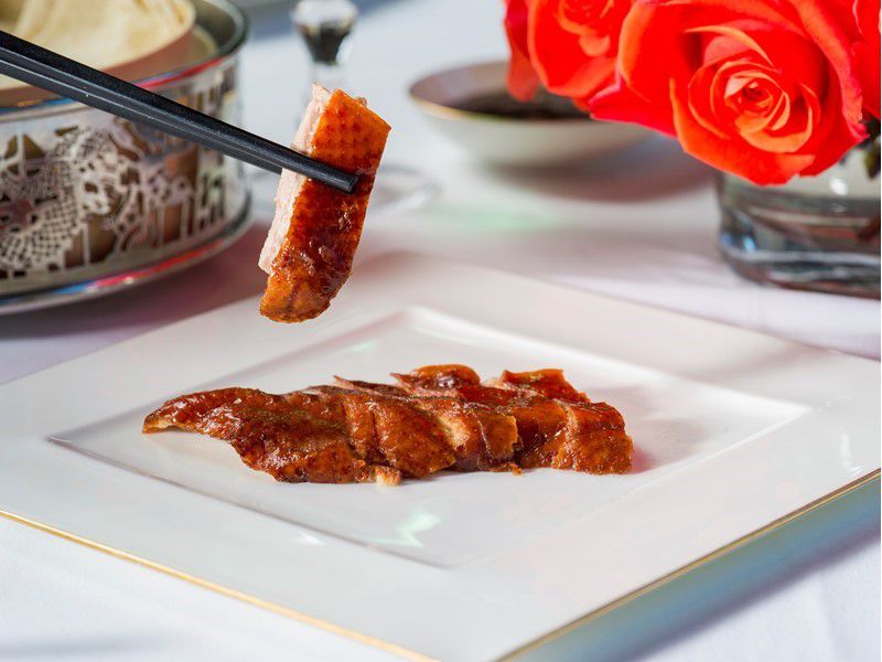 A square white plate with a few pieces of Peking duck, one held aloft with chopsticks, in front of red roses.&nbsp;