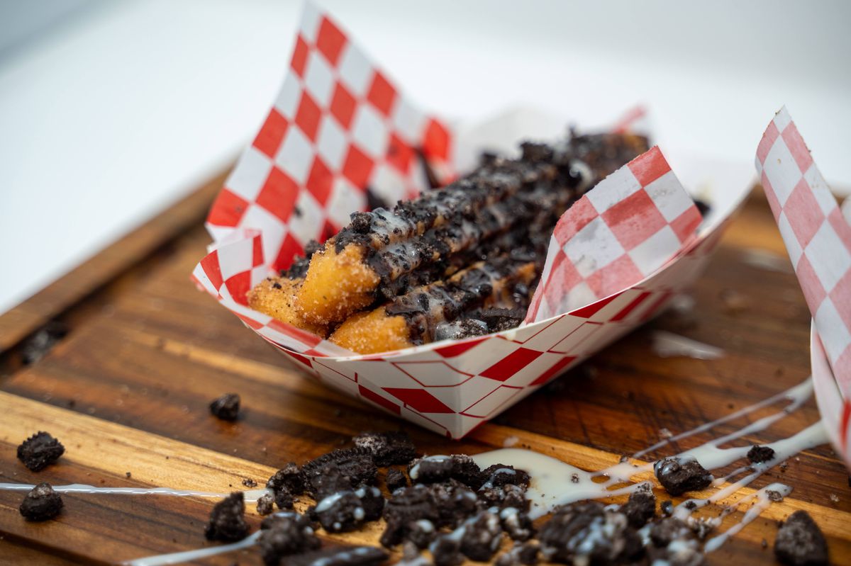 A cardboard boat with a red-checked paper holds churros coated with chocolate syrup, cream cheese icing, and smashed Oreos.