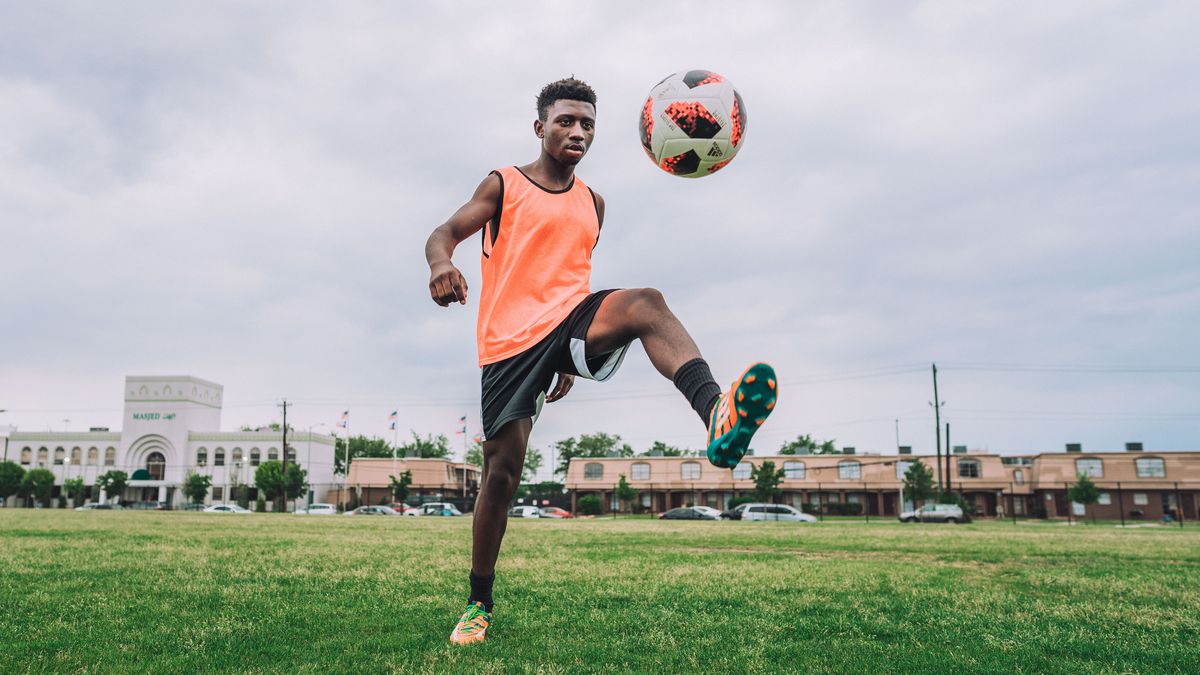 A refugee soccer player in an orange tank top juggling a soccer ball on an empty field in Houston.