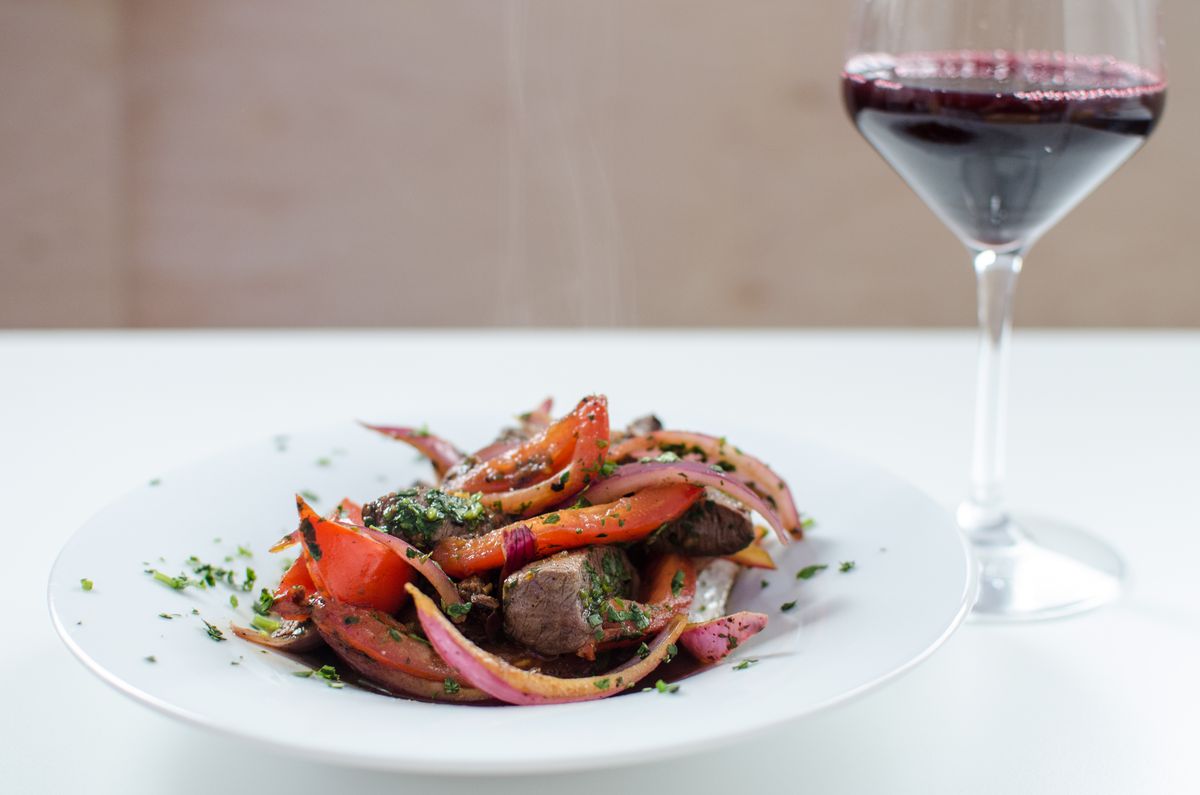 A white plate sits on a white table, full of cubes of marintaed beef with peppers and tomatoes. A glass of red wine sits to the right.