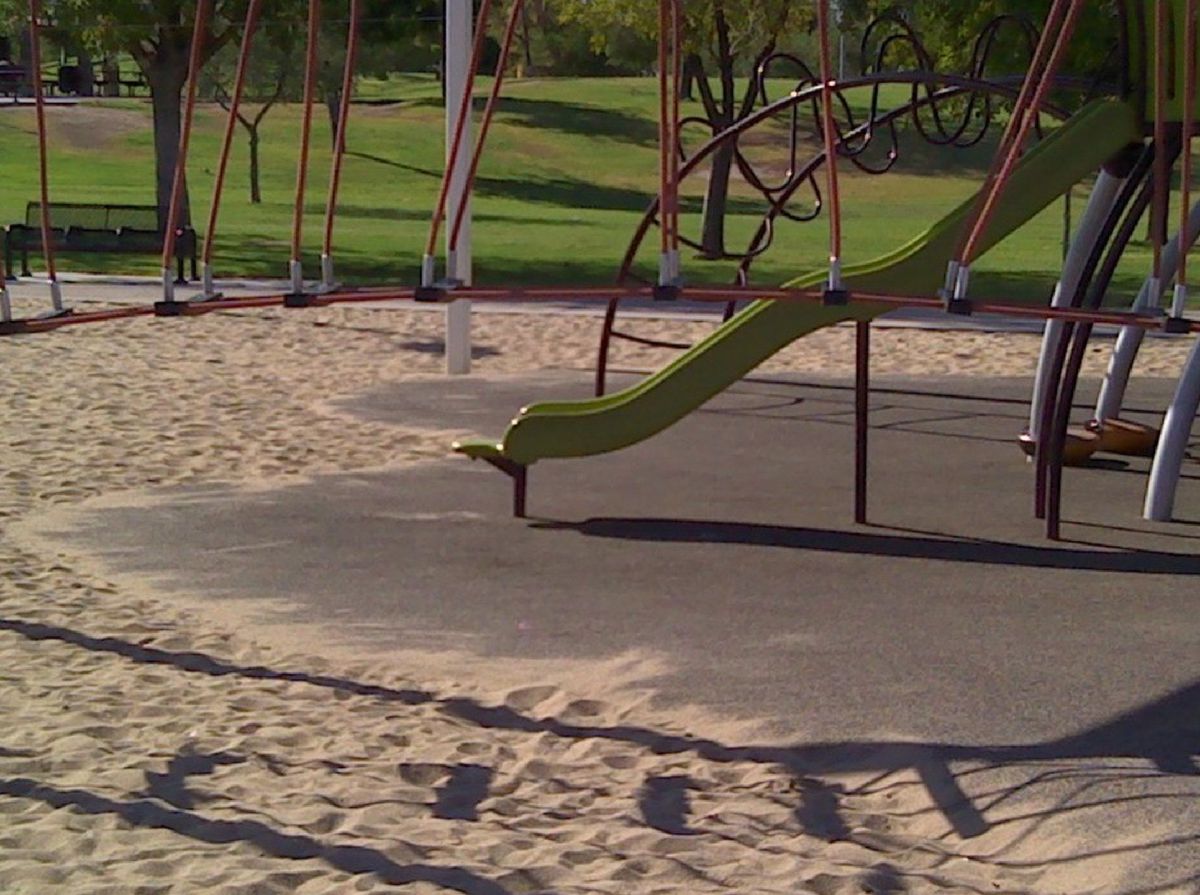 The green slide at a sunny playground is surrounded by other playground equipment and the grass of a park in the background.