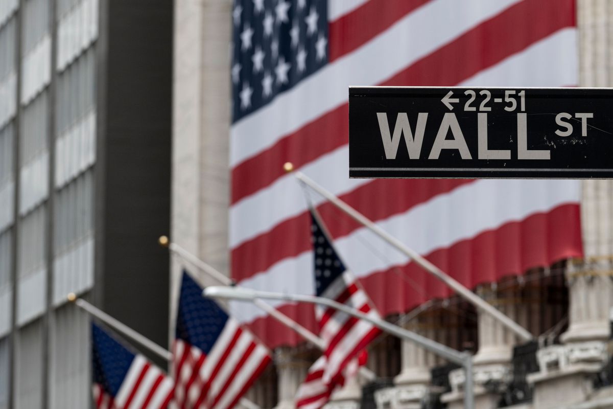 A Wall Street sign with the New York Stock Exchange building behind it displaying American flags.