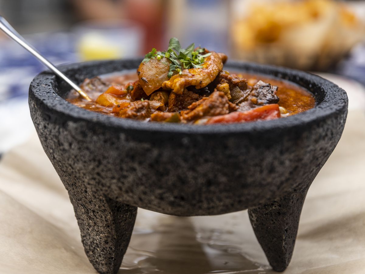The main focus of this picture is a lava bowl filled with Carne Molcajete. A dish served at Rocco’s Tacos that is beef stewed in tomato sauce and peppers. There are some blue plates and tortilla chips that are out of focus in the background.