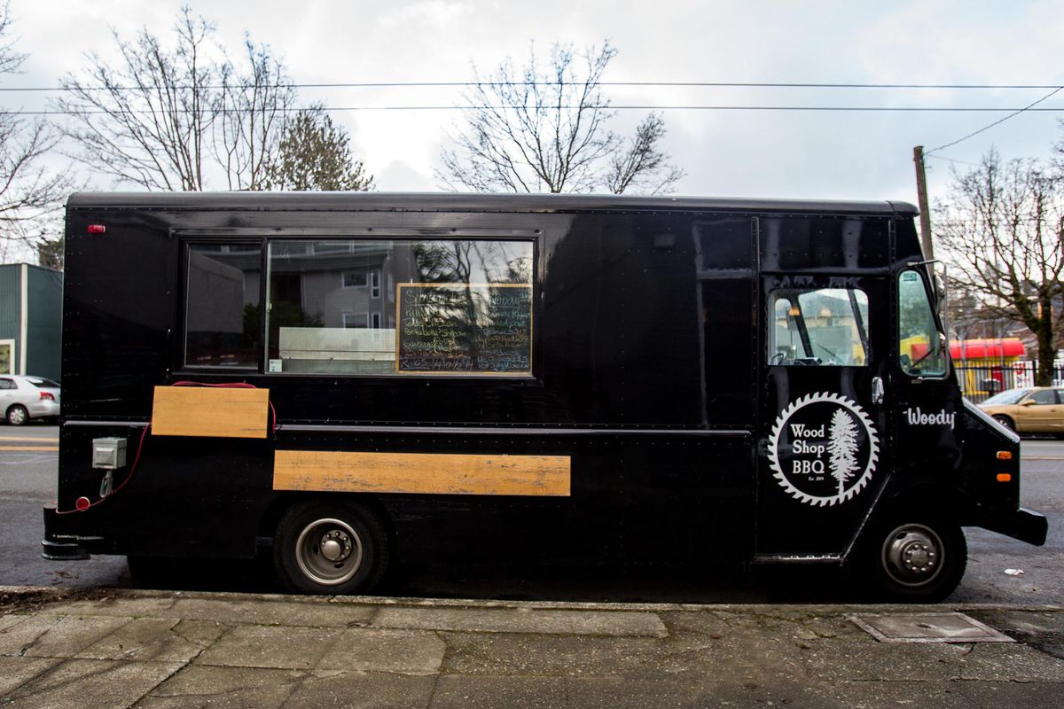 The side view of a black food truck with the Wood Shop BBQ logo in white on the door.
