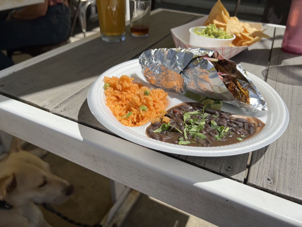 a taco plate with beans and rice on a picnic table. Below, a dog sits patiently.