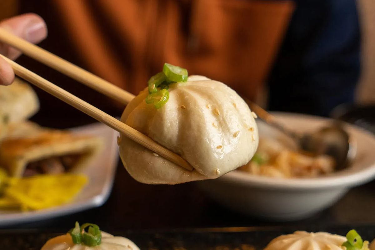 A soup dumpling grasped by chopsticks with other dishes blurred in the background.