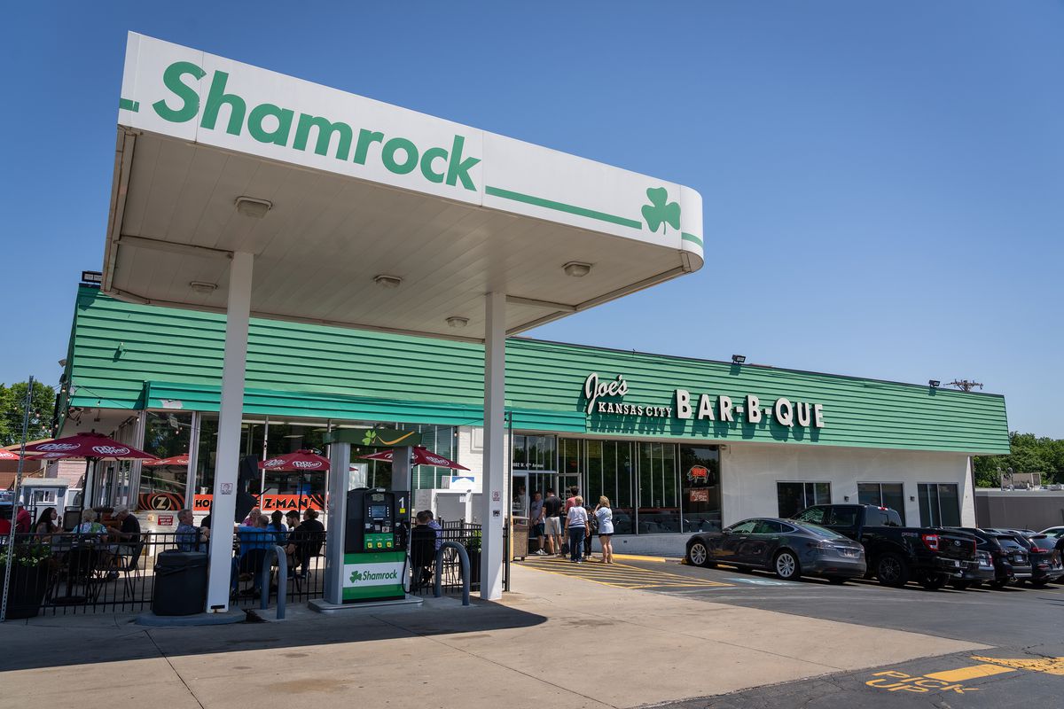 A restaurant exterior set into a Shamrock gas station. The restaurant name reads Joe’s Kansas City Bar-B-Que. Diners wait in a line outside. 