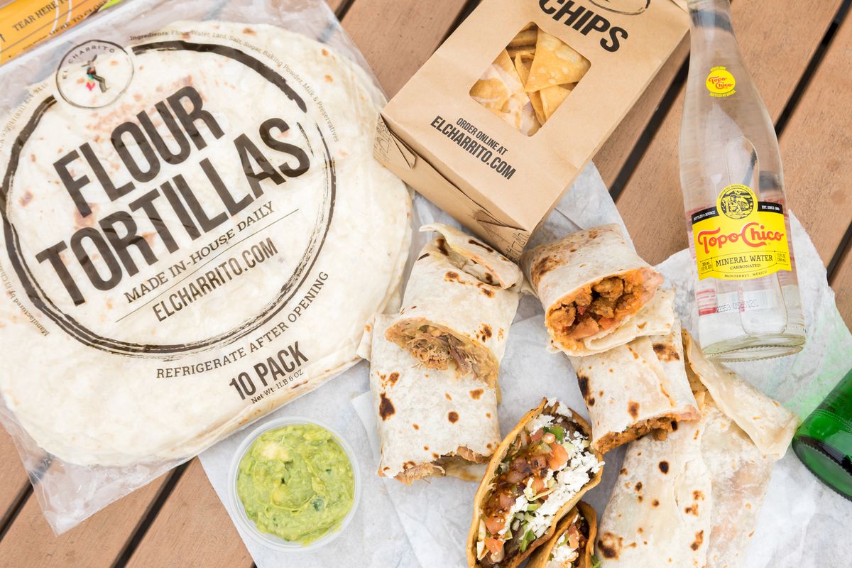 A pack of 10 flour tortillas, guacamole, tortilla chips, burritos, and a Topo Chico bottle from El Charrito are displayed on a table.