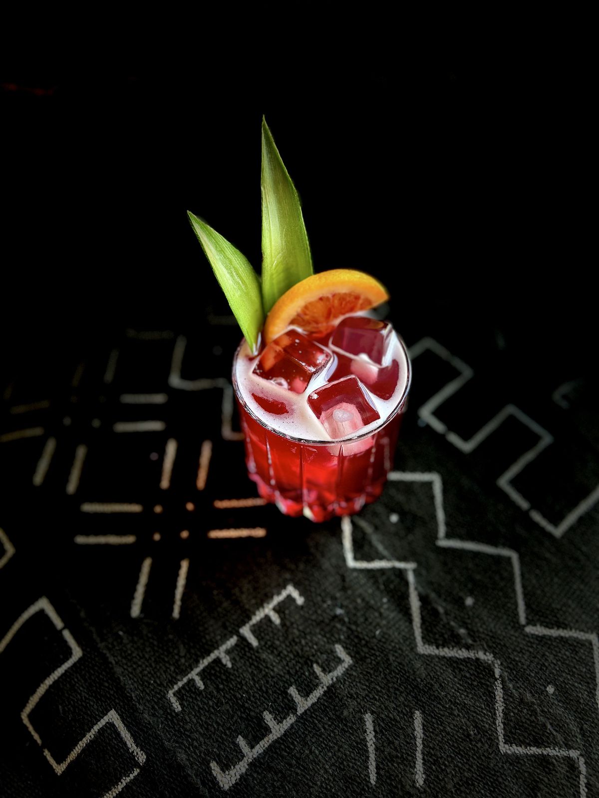 A red cocktail with large ice cubes, two green pointy leaves, and a grapefruit slice on a black patterned surface.