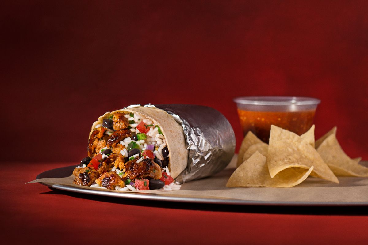 A Chipotle chorizo burrito next to a bag of chips and a plastic container of salsa, up against a red backdrop.