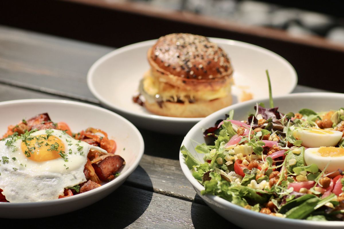 Three plates sitting on a dark wood table. one plate features potatoes with fried egg on top, a second plate features a salad, and a third plate features a breakfast sandwich with egg, cheese, and a poppy seed bun.