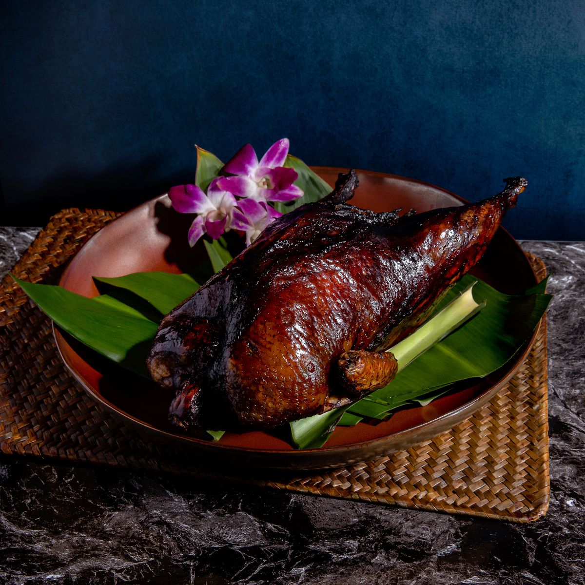 A roasted whole duck sits on banana leaves on a bamboo tray; it’s garnished with orchids.