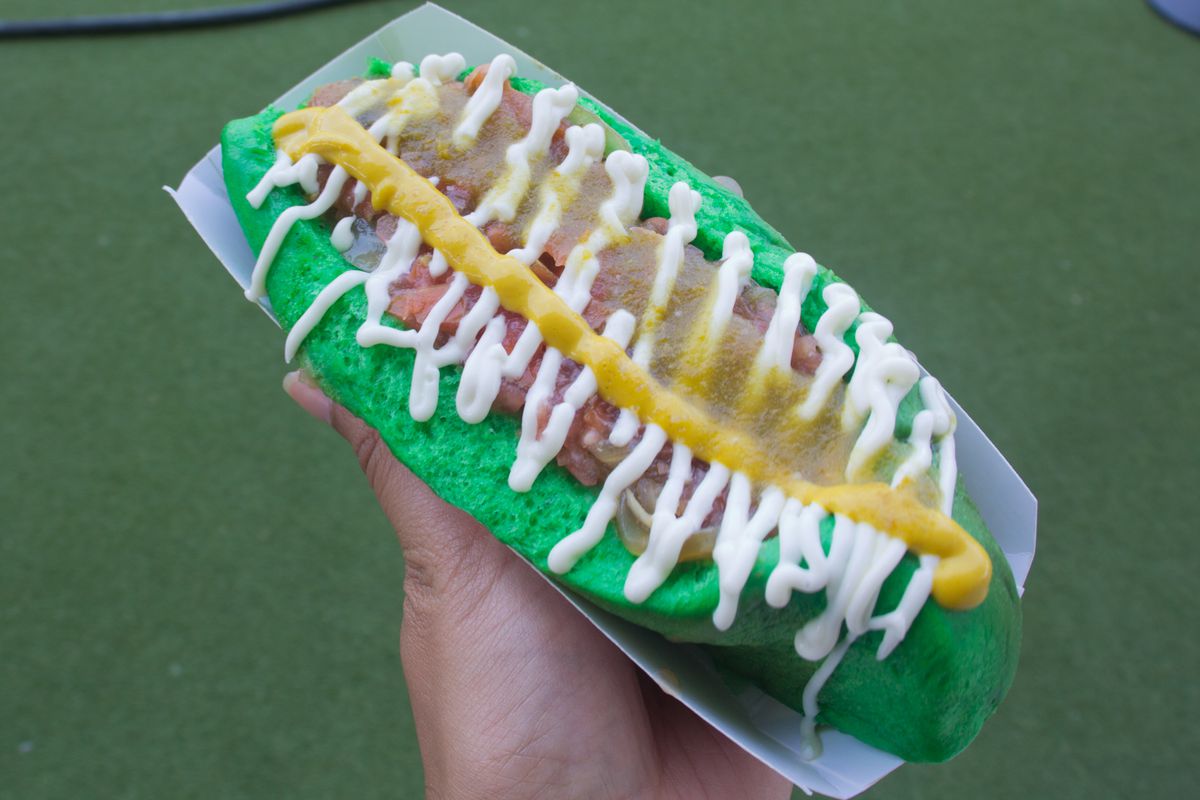 A green hot dog bun with a hot dog and layers of drizzled sauces in white, green, and red