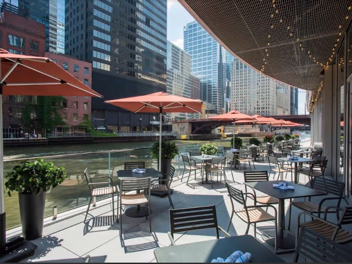 A patio along a river with covered tables in the foreground and skyscrapers in the back.