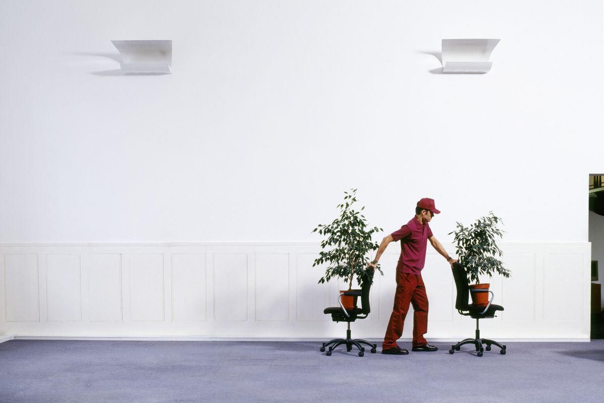 A photo of a man in a worker’s uniform wheeling office chairs, on top of which plants perch, through an empty-looking office setting.