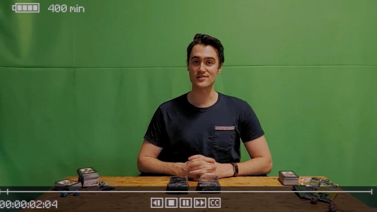 Inscryption - Yeşil ekran arka planının önünde genç bir adam olan Luke Carder oturuyor ve kart oyunları hakkında bir video kaydetmeye hazırlanıyor