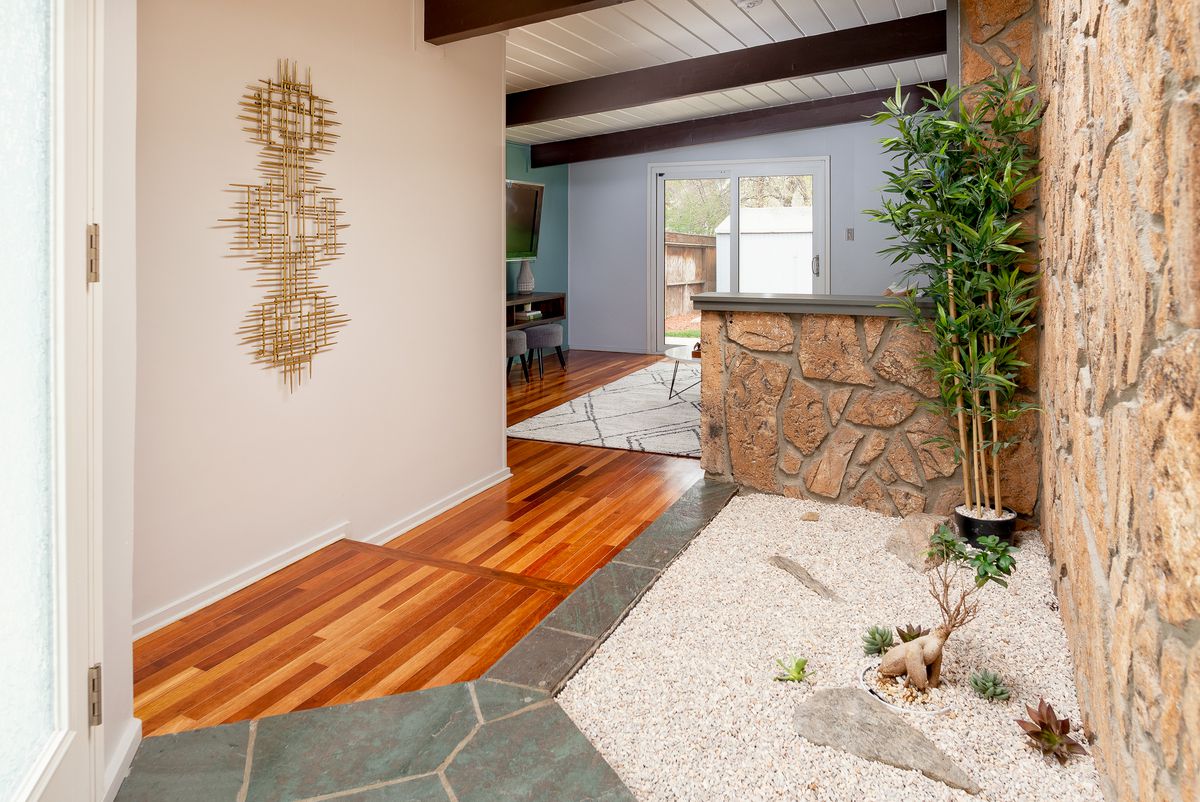 The interior entrance to the ranch home has wood floors and a small planter area with bamboo, white rock, and a wall of moss rock. 