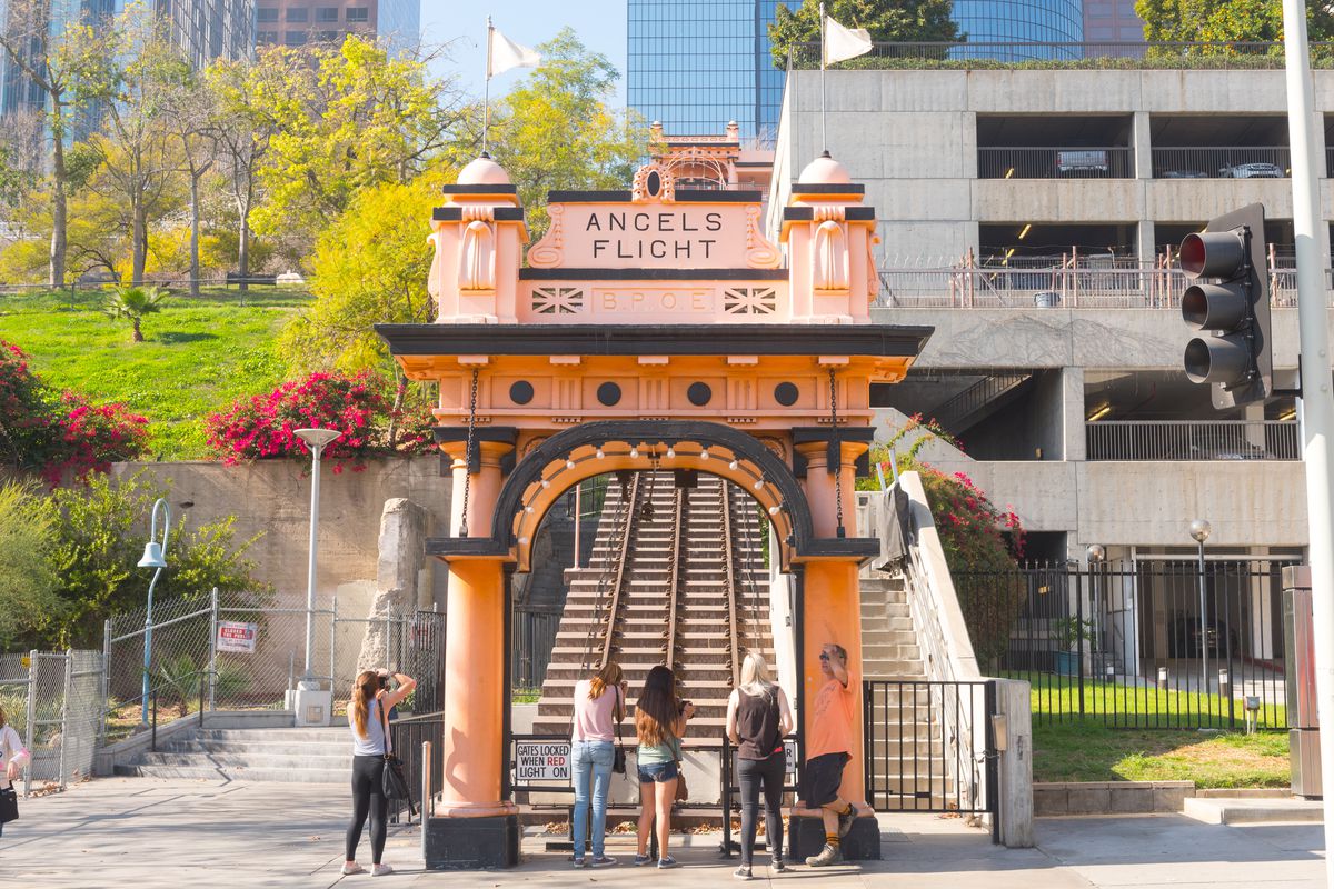 In the foreground is the entrance to a railway with train tracks. The sign on the entrance reads Angels Flight. Behind the railway are buildings and a park with trees and flowers.