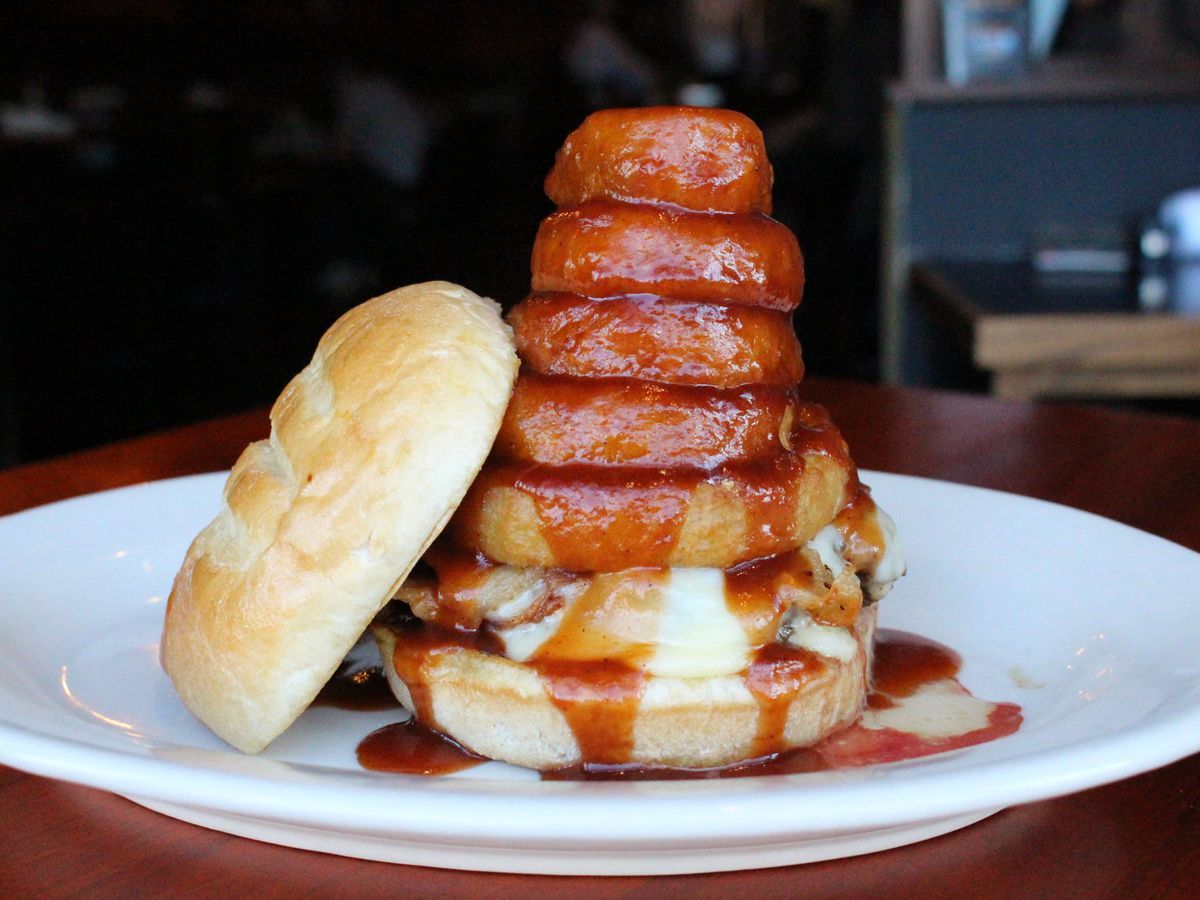 A burger sits on a white plate, its top bun leaning up against it. It’s topped with a stack of five thick onion rings that are drenched in barbecue sauce.