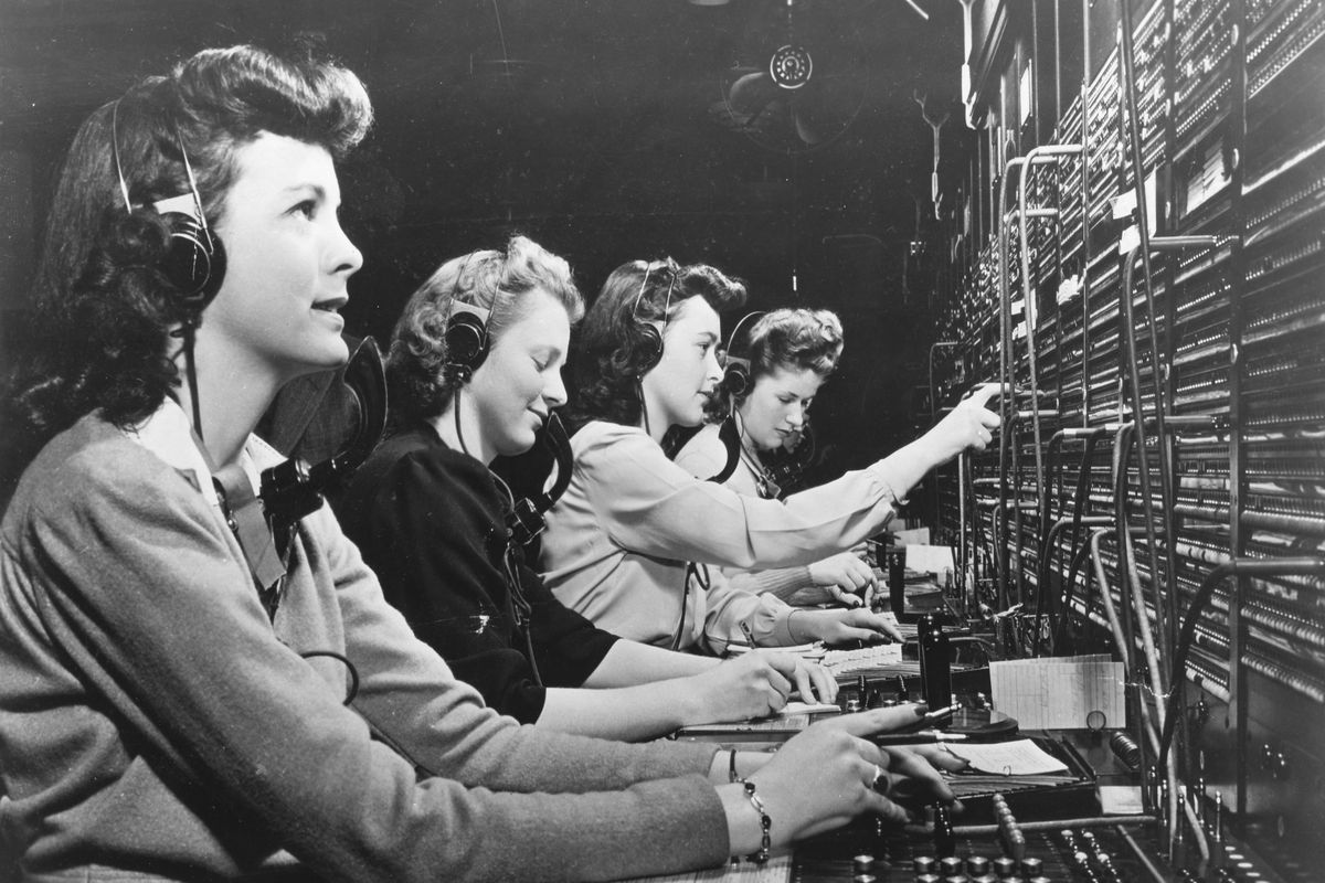 A black-and-white photo shows a row of young women in 1940s fashion working at a telephone switchboard.