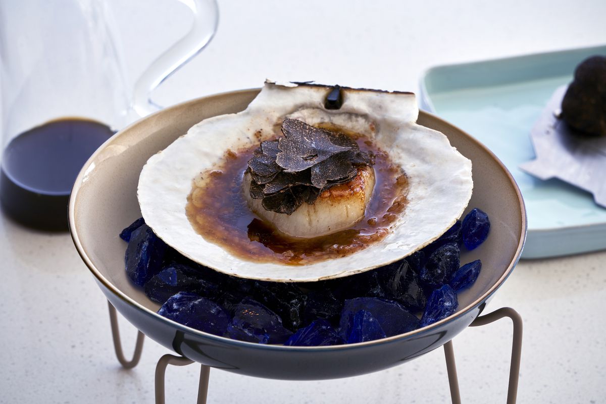 A side angle shot of a scallop in a large shell on top of blue rocks in a dark bowl.