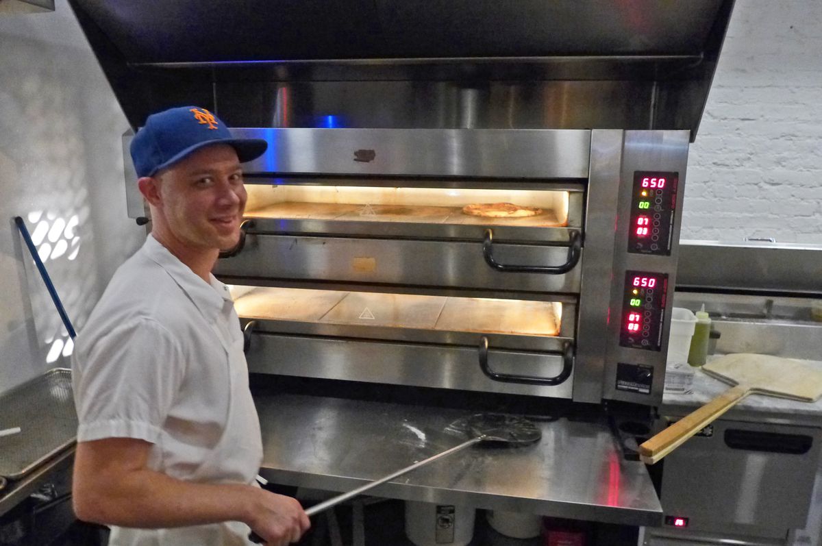 A man with a wooden pizza paddle stands in front of double stacked ovens.