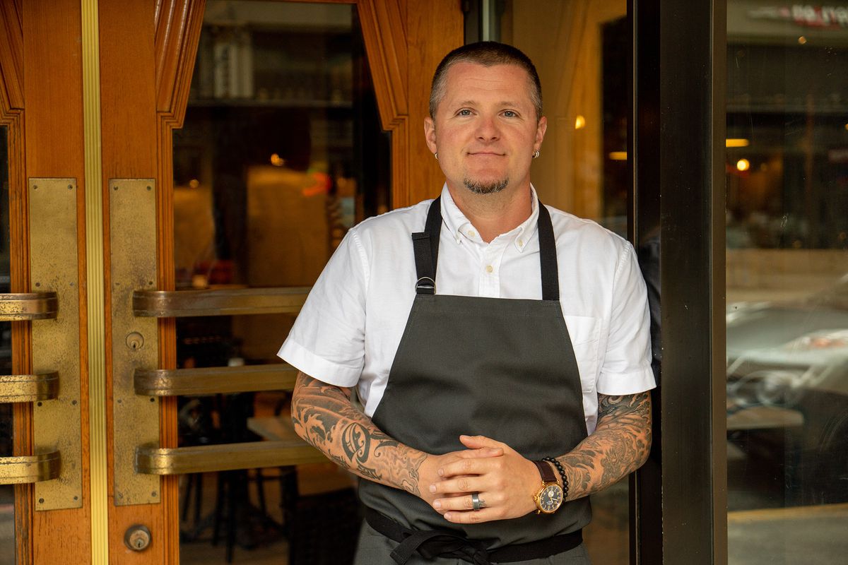 A man wearing a black apron and a short-sleeved white shirt with tattoos and a buzz cut stands in front of the wooden doors of a restaurant. 