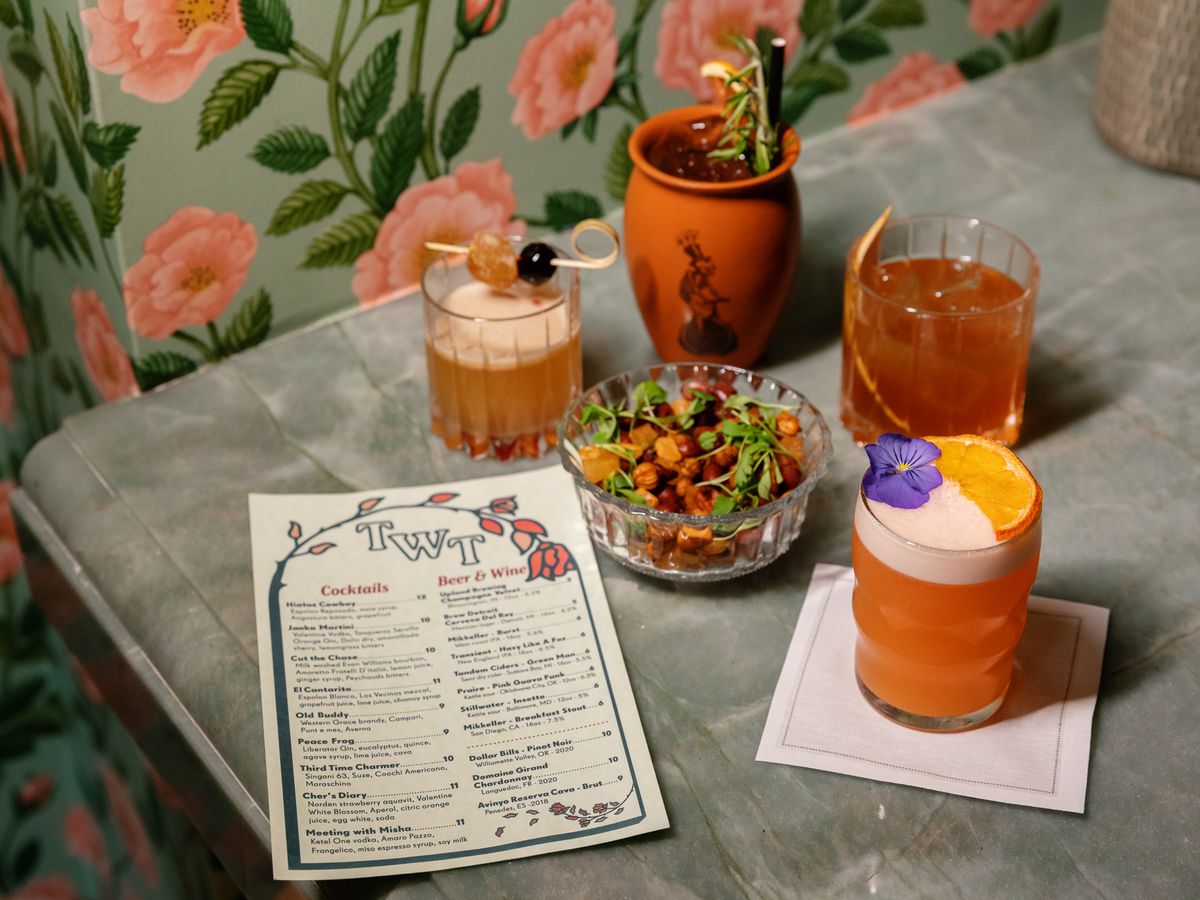 Four drinks, one in a clay jar, three in glass, one set on top of a napkin, a bowl of mixed nuts, a menu all set on top of a white stone top table and floral wallpaper in the background