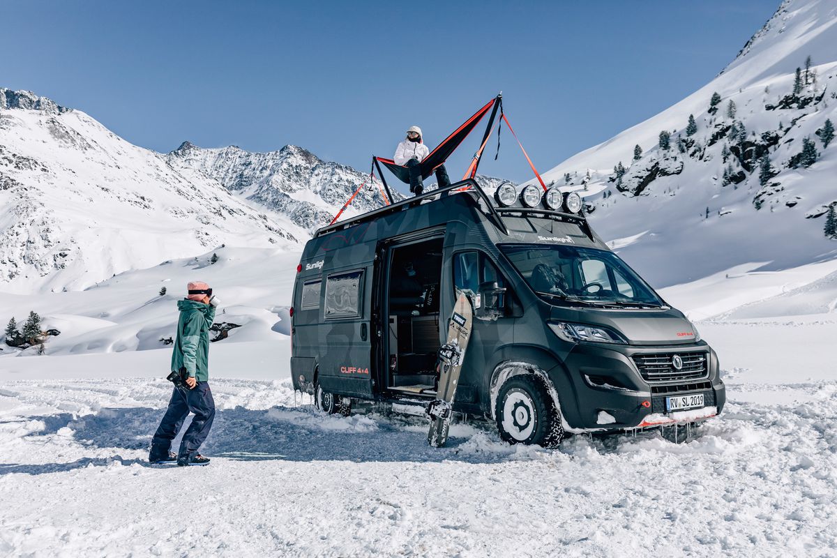 A gray camper van sits in a snowy valley below mountains and a blue sky. The side door of the van is open with a snowboard standing next to it; one person in snow gear is walking towards the van and another person sits on the roof in a hammock.