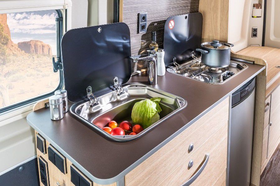 A kitchenette inside of a RV camper van. The counter has a compartment which is open and has fresh produce in it. There is another compartment with a stove that has a large pot on it. 