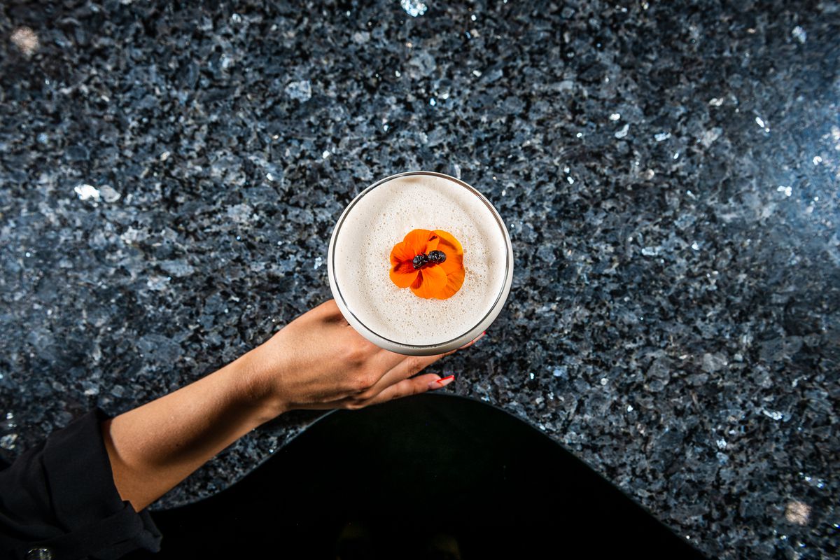 A black aunt sits on an orange flower floating on the top of a frothy Chicatana Sour cocktail