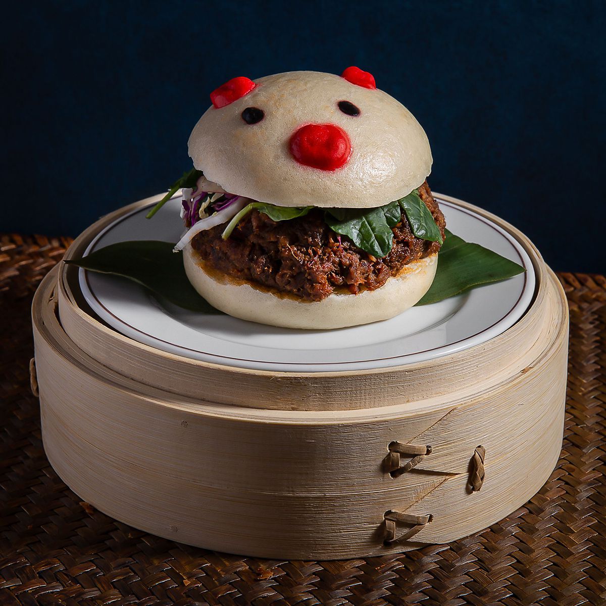 A pork bao sits on a steamer basket. The bao bun is pig-shaped and decorated.