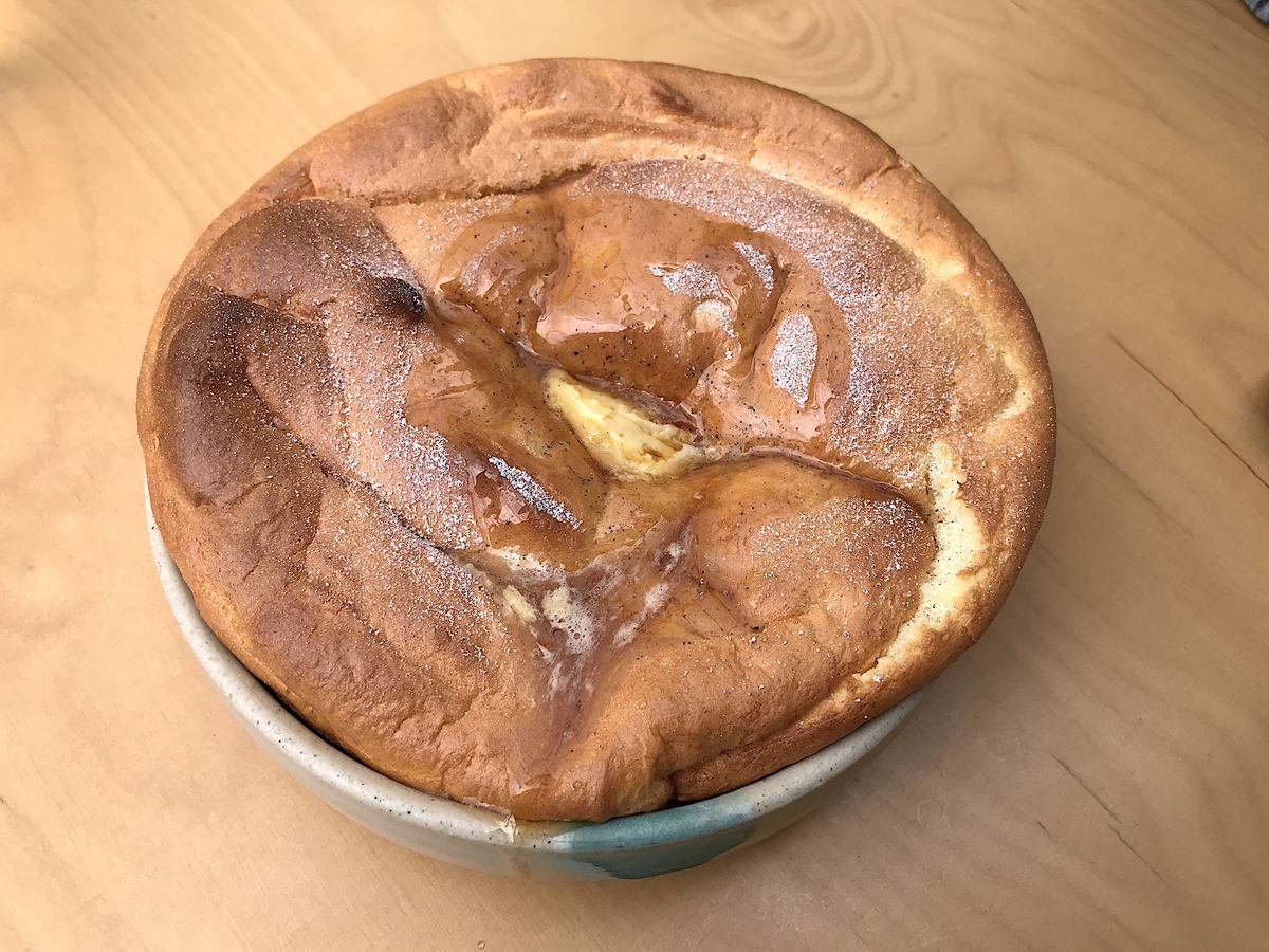A deflated but golden brown souffle rests in a tin dish on top of a wooden table