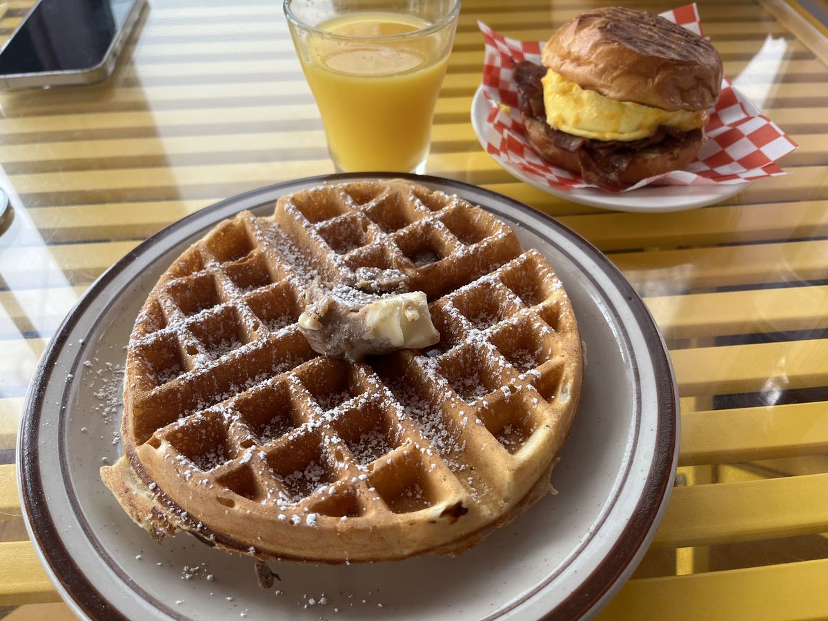 A waffle with a dab of butter and a powdered sugar topping next to a breakfast sandwich.