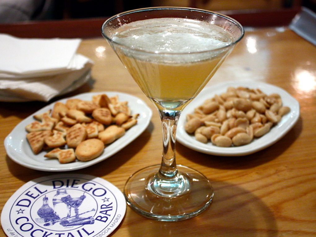 A cocktail in a martini glass sits in the middle of several small plates of crunchy snacks and a coaster branded with the logo of Del Diego Cocktail Bar.