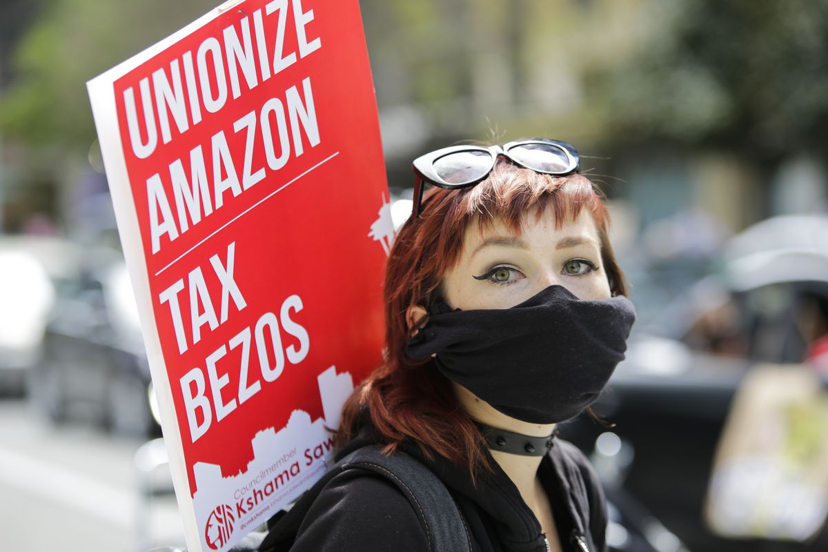 A protester in a breathing mask carries a sign reading “Unionize Amazon, tax Bezos.”