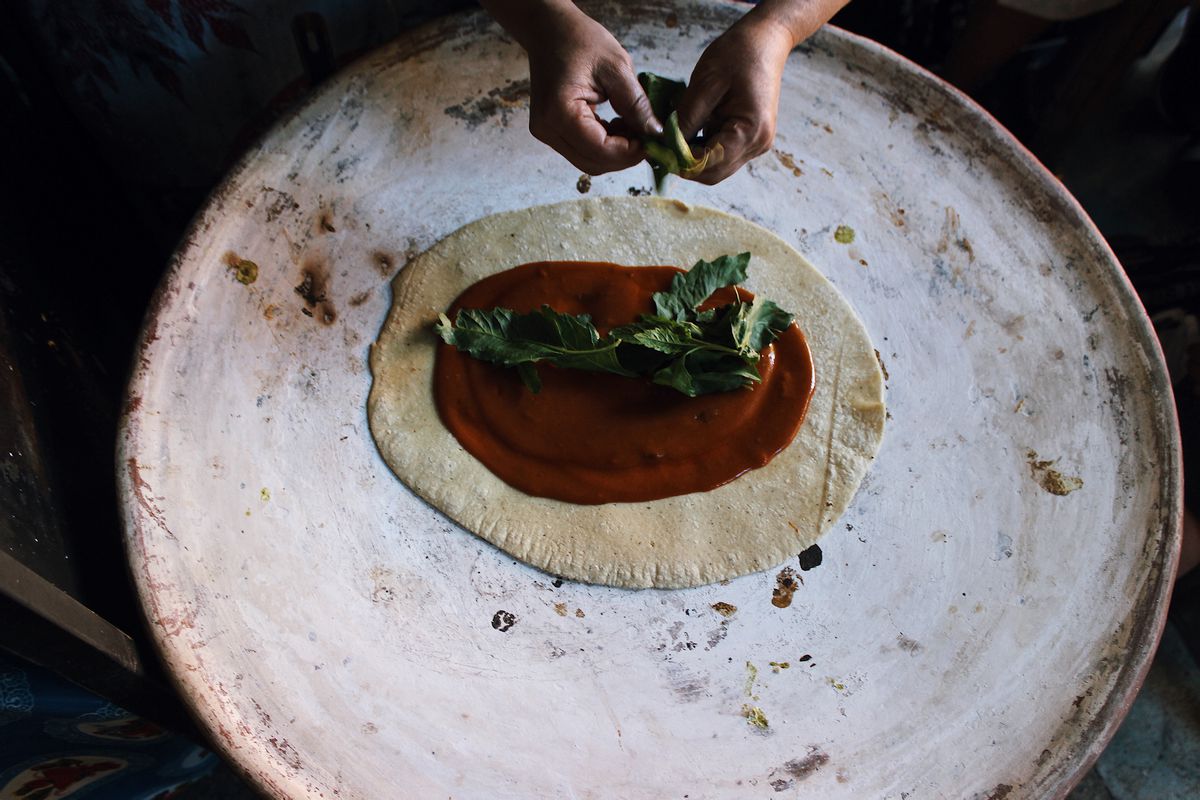 A large round of masa has red sauce and herbs, cooking on a comal. 
